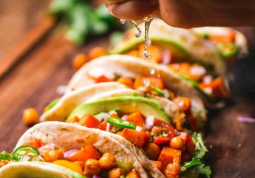 The image shows several tacos filled with chickpeas, vegetables, and avocado slices, with a hand squeezing lime juice over them.