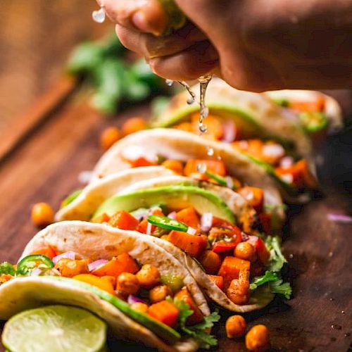 The image shows several tacos filled with chickpeas, vegetables, and avocado slices, with a hand squeezing lime juice over them.