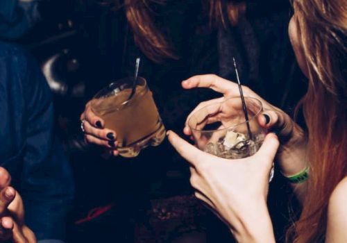 The image shows a group of people at a social event, holding drinks and engaging in conversation, with one person laughing.