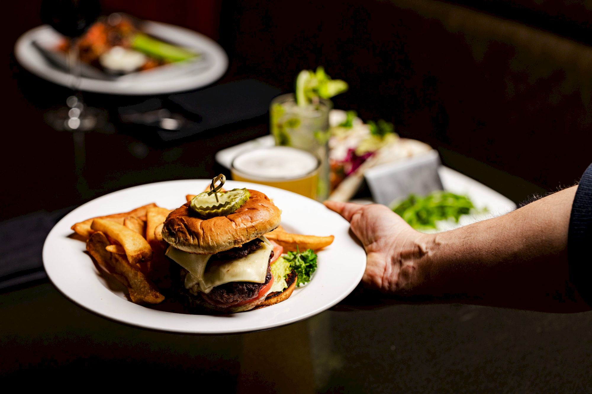 A plate with a cheeseburger, fries, and garnish is being served, with other food and drinks visible in the background.