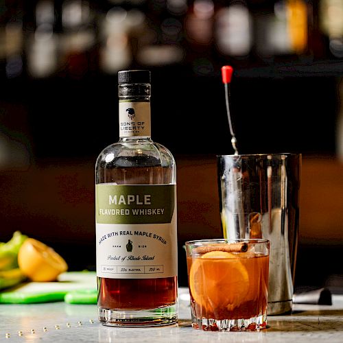 A bottle of Maple Bourbon Whiskey, a cocktail mixing tin, and a glass with an orange garnish are on a bar counter.