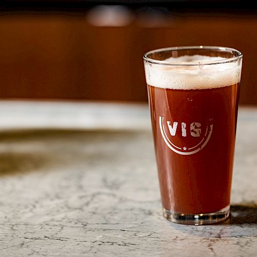 A pint glass filled with amber beer, labeled "VIS," sits on a marble surface.