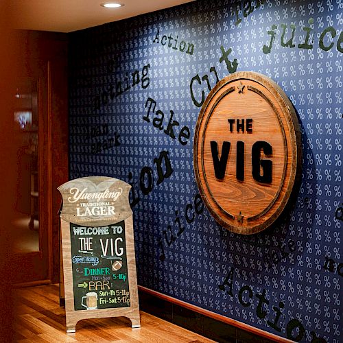 The image shows a wall with a circular wooden sign reading "THE VIG" and a smaller sign below with "WELCOME TO THE VIG" and other text.