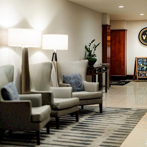 A lobby with comfortable chairs, lamps, a rug, a potted plant, and a sign in the background showing the word 