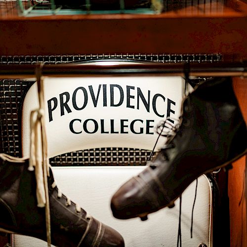 The image shows a display with a sign that reads "Providence College" and two old-fashioned football shoes hanging above it, suspended from a rod.