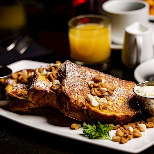 The image shows a plate of French toast topped with nuts, served with butter, syrup, and garnished with parsley. There’s also coffee and orange juice.