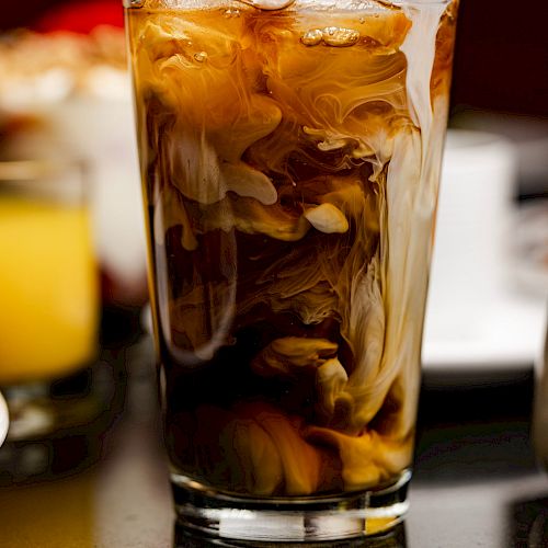 The image shows a glass of iced coffee with cream swirling through it, placed on a reflective surface.