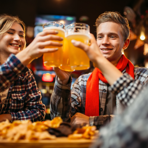 Three people are raising glasses of beer in a joyful toast at a bar, with snacks in the foreground, and festive lights in the background.