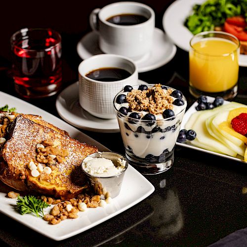 A breakfast spread with French toast, yogurt with blueberries and granola, a plate of sliced fruits, coffee, orange juice, and a salad.