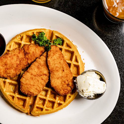 A white plate of chicken and waffles with syrup and whipped cream on the side, garnished with parsley, placed on a dark surface.