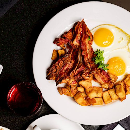 The image shows a plate with bacon, fried eggs, and diced potatoes, alongside a small bowl of fruit and a glass of red liquid.