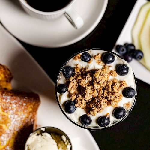 A breakfast spread with yogurt and granola, fresh fruits, coffee, French toast with nuts and whipped cream on the side, and melon slices.