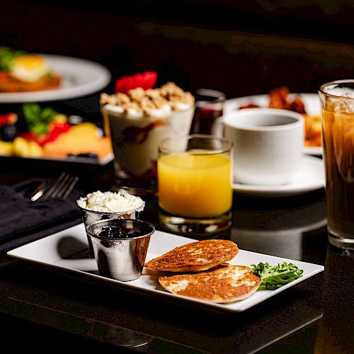 A breakfast spread featuring pancakes, fruit, yogurt parfaits, orange juice, iced coffee, and other breakfast items on a black table ending the sentence.