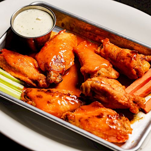 A tray of buffalo chicken wings with dipping sauce, celery sticks, and carrot sticks, served on a white plate.