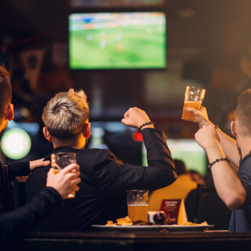 Three people are in a bar, watching a soccer game on TV and cheering while holding drinks, with food on the table in front of them.