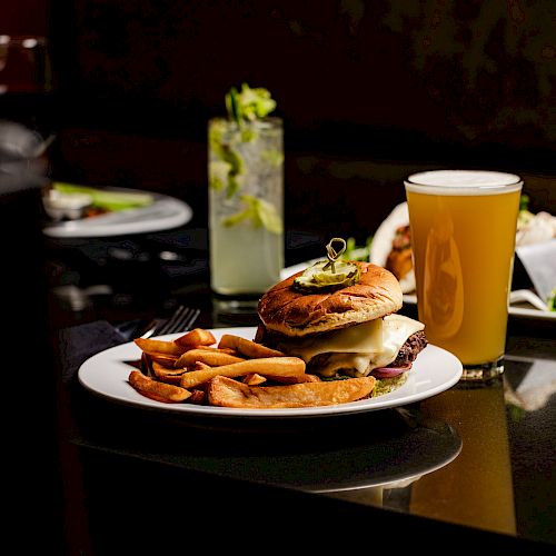 A plate with a burger and fries, a glass of beer, a cocktail, and some greens on a table in a restaurant setting.