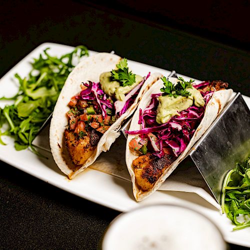 The image shows two tacos with grilled filling, red cabbage slaw, and a green sauce on a white plate, garnished with greens, next to drinks.