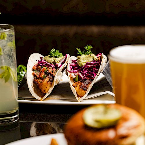 The image shows a plate with two tacos, a glass of light beer, a cocktail with mint garnish, and part of a sandwich at a dining table.