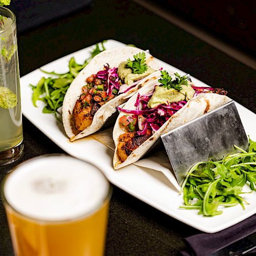 The image shows two tacos filled with vegetables and garnished with greens on a plate, accompanied by a drink, possibly beer, in the foreground.