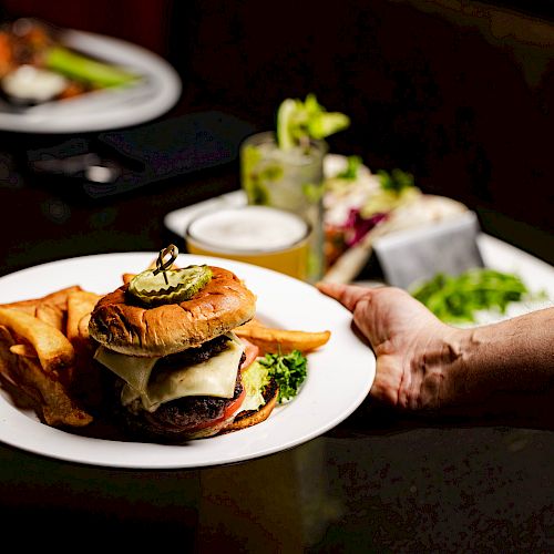 A hand is holding a plate with a burger and fries. A table with drinks, a salad, and another plate are in the background.