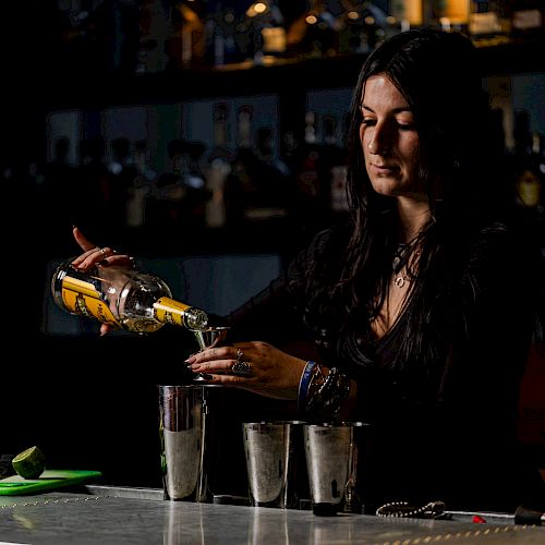 A person is behind a bar pouring a beverage into three metal shakers, with various bottles visible in the background, suggesting a bar setting.