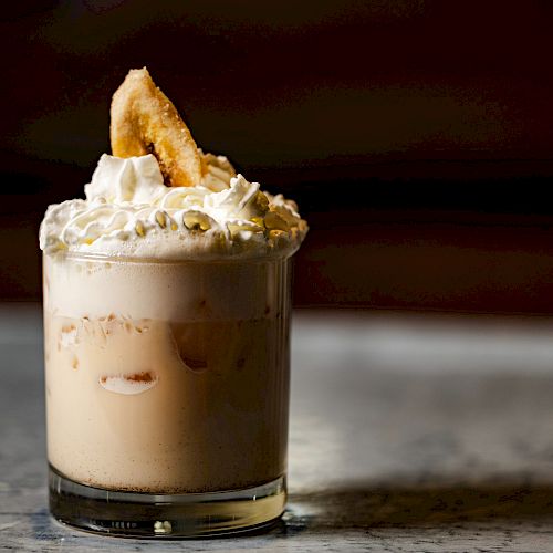 A creamy beverage in a short glass with whipped cream and a banana slice on top, set against a dark background on a marble surface.