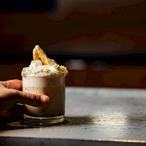 A hand holding a dessert drink topped with whipped cream and a slice of fruit, set on a marble surface.