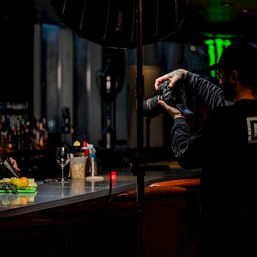 A photographer is capturing a picture of a bartender preparing a drink at a well-lit bar, with various bar tools and ingredients on the counter.