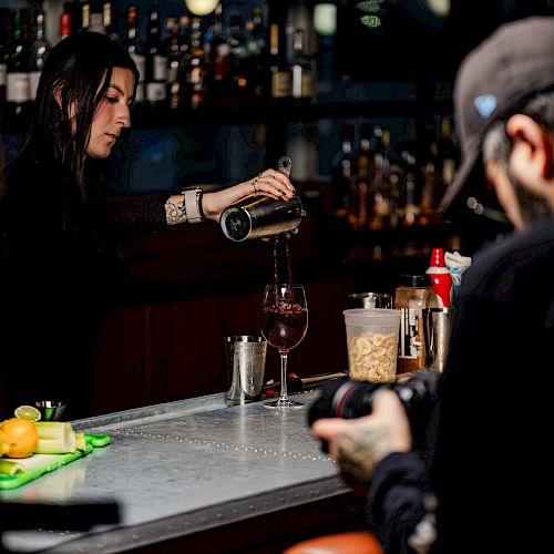 A bartender is pouring a drink into a glass behind a bar, with another person in the foreground seemingly taking photos.