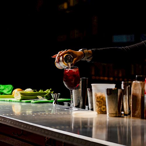 A hand pours a drink from a can into a glass on a bar counter, surrounded by bar tools and ingredients under dim lighting.