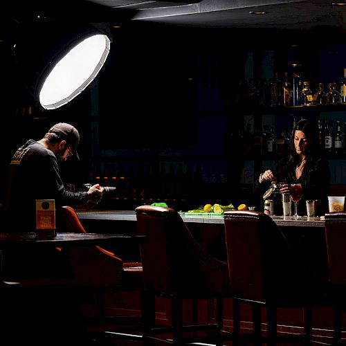 A person is seated at a dimly lit bar counter with a bright light overhead, while another person stands behind the bar counter.