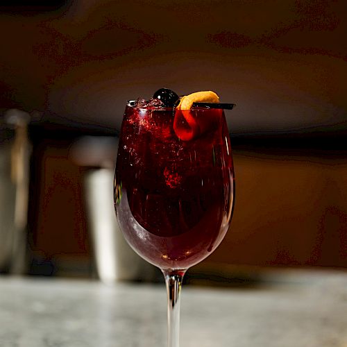 A glass of red sangria with fruit garnishes sits on a bar counter, featuring an elegant stem and a dark, blurred background.