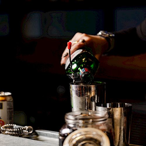 A person is pouring liquid from a green bottle into a metal container on a bar counter, surrounded by jars and metal shakers.