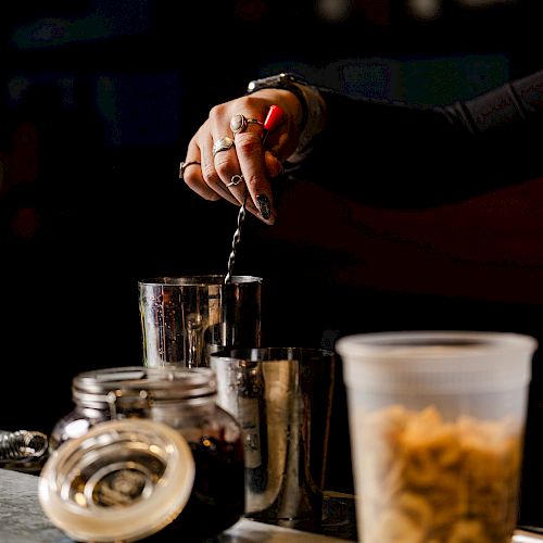 A person is mixing a drink in a shaker behind a bar, with various containers and a can nearby on the counter.