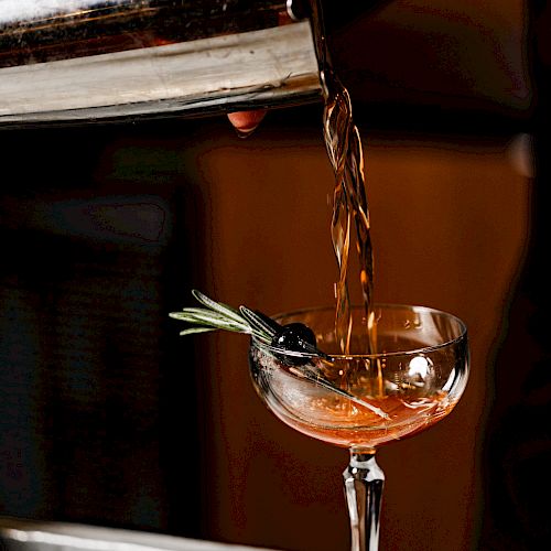 A cocktail is being poured from a shaker into a coupe glass, garnished with herbs and a cherry on a pick.