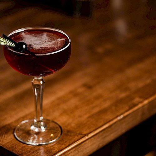 A cocktail in a coupe glass, garnished with a cherry and a sprig of herbs, sits on a wooden surface.