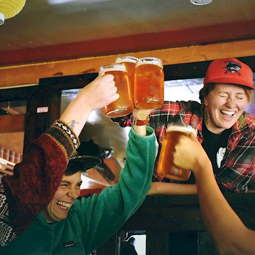 Four friends are raising beer mugs and cheering, smiling and laughing in a warm, indoor setting with colorful hanging lights.