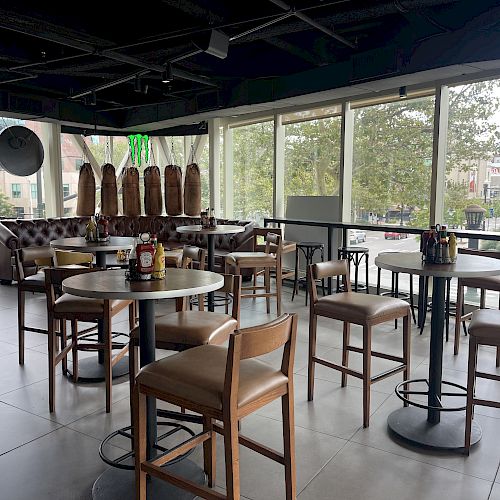 A modern indoor dining area with wooden tables and chairs, condiment bottles, and large windows offering a view of trees and buildings outside.