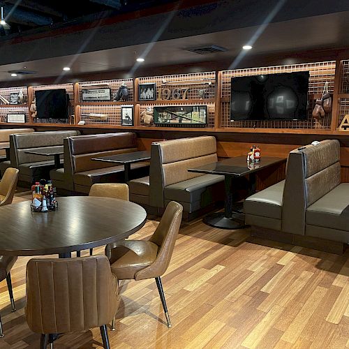 The image depicts a modern, cozy restaurant or cafe interior with wooden floors, booth seating, round tables, and wall decor, including TV screens.