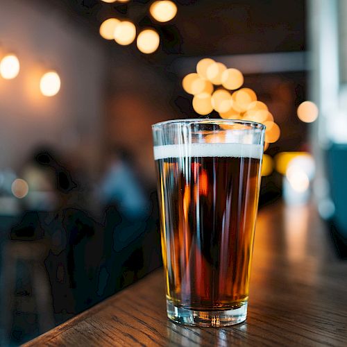 A glass of beer is placed on a wooden surface in a dimly lit environment, with blurred lights and people in the background.