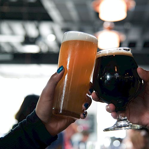 Two people are clinking glasses of beer, one with a light amber color and the other with a dark, rich hue, in a social setting.