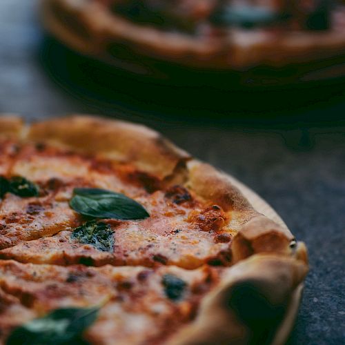 The image shows two delicious-looking pizzas with a golden-brown crust and toppings of basil leaves on a dark surface.