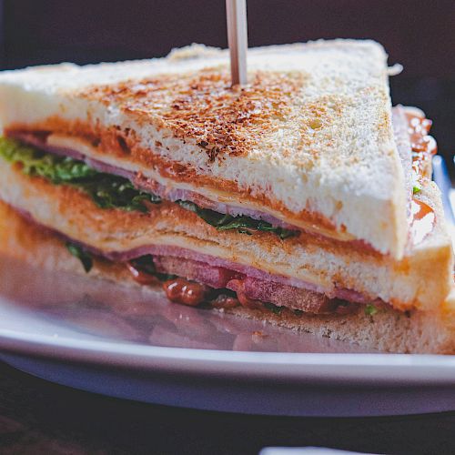 A close-up of a club sandwich on a white plate with layers of bread, lettuce, tomato, and meat, accompanied by a napkin.