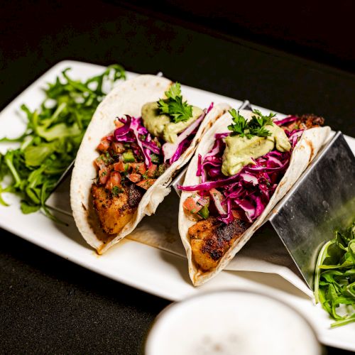 The image shows two tacos filled with grilled meat, red cabbage, and avocado, garnished with greens, served on a white plate.