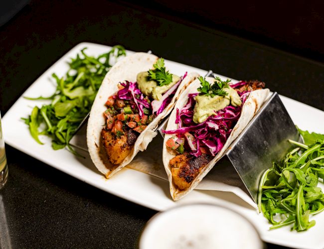 The image shows two tacos filled with grilled meat, red cabbage, and avocado, garnished with greens, served on a white plate.