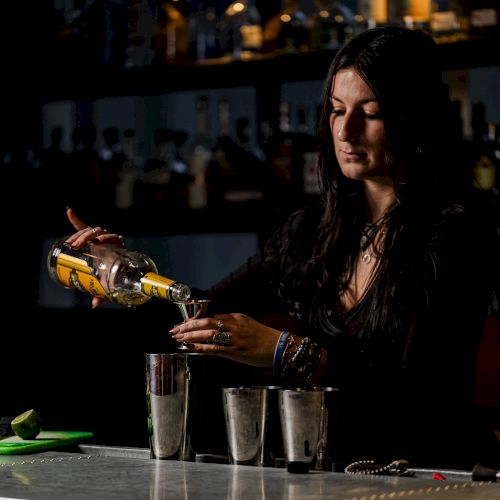 A person behind a bar counter pouring a drink from a bottle into three metal cups. The background showcases bottled alcohol on shelves.