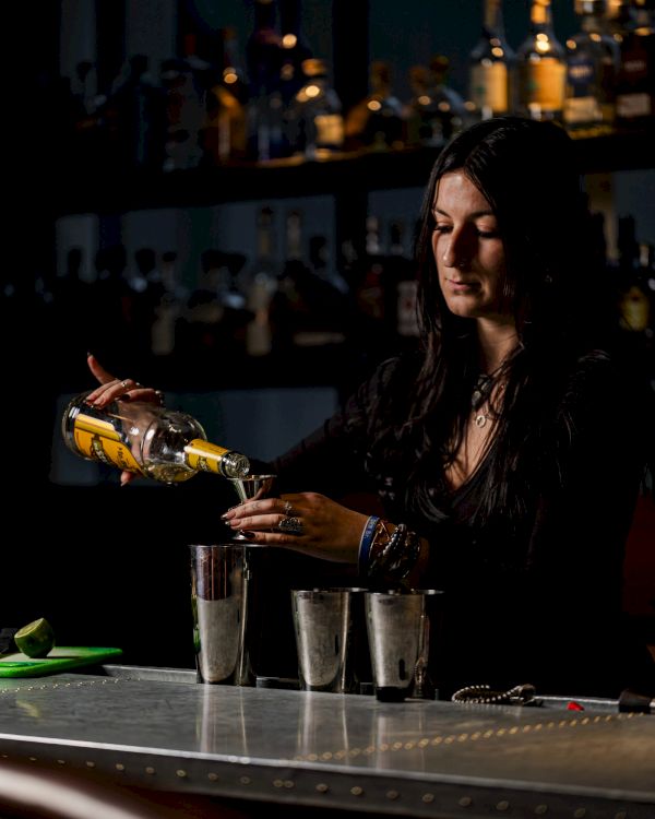 A person behind a bar counter pouring a drink from a bottle into three metal cups. The background showcases bottled alcohol on shelves.