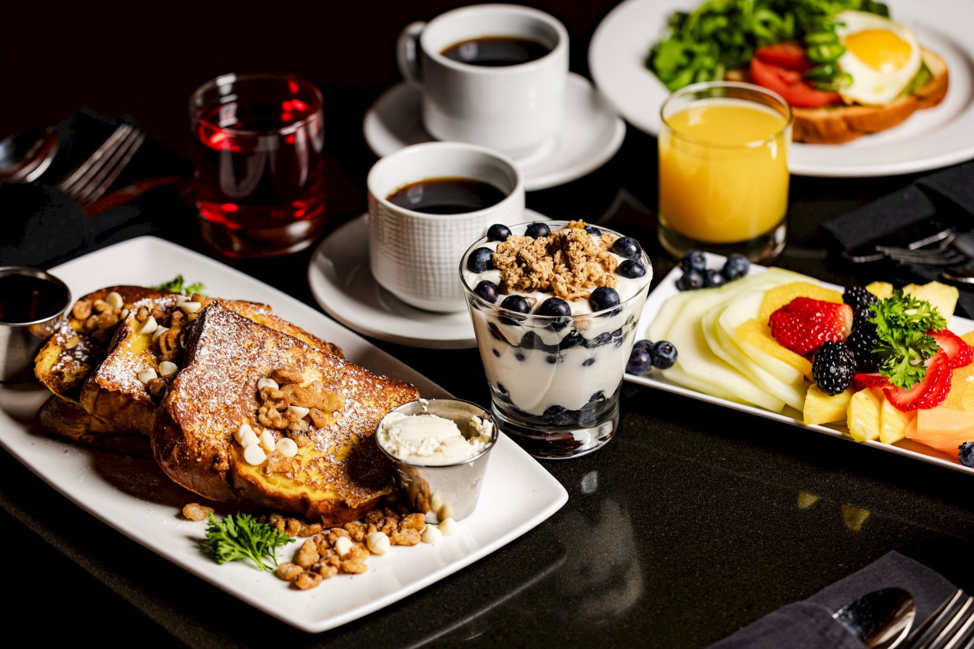 The image shows a breakfast spread with French toast, granola with yogurt and blueberries, fruit salad, coffee, orange juice, and tea.