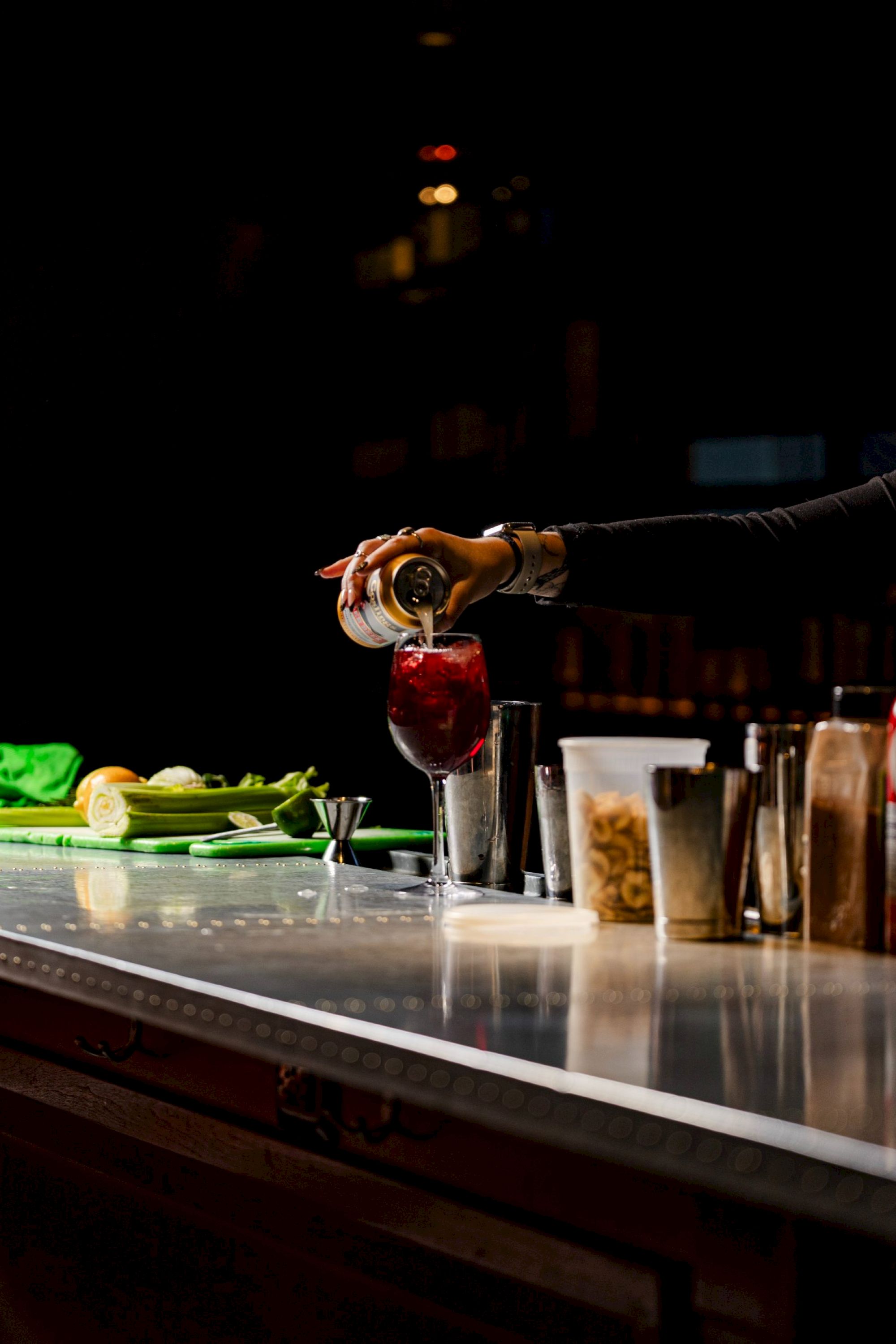 A person is pouring a drink into a glass at a bar counter with various ingredients and utensils around.