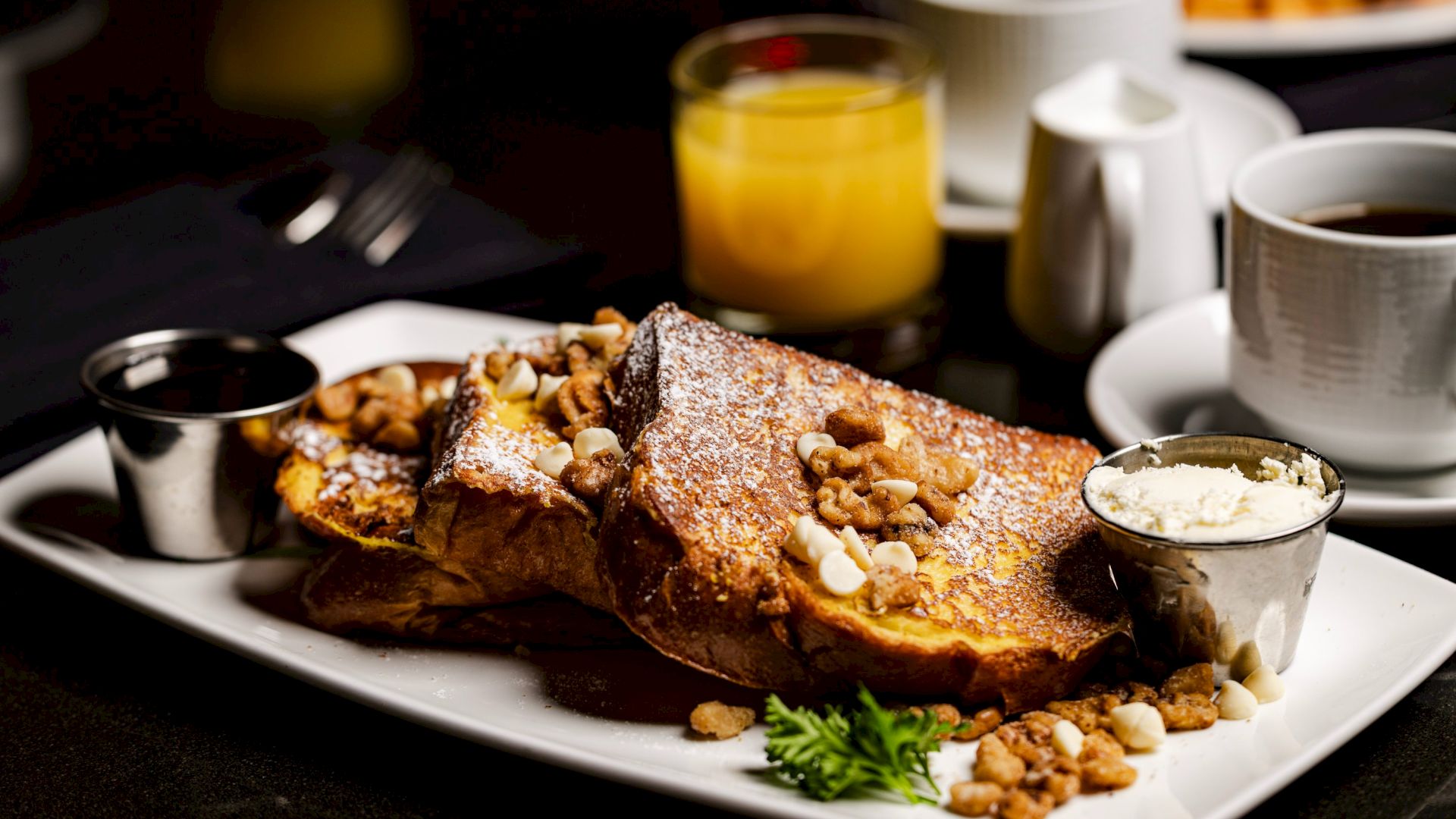 The image shows a delicious plate of French toast topped with nuts, served with a side of cream, syrup, orange juice, and a cup of coffee.